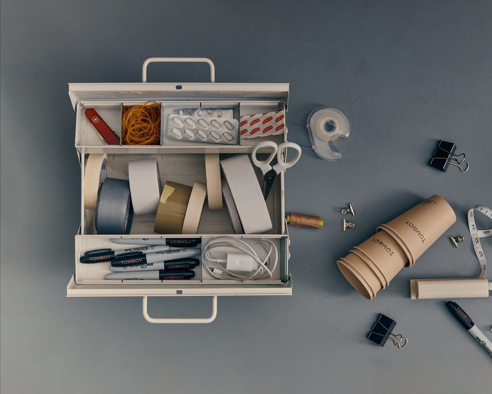 Tomboy Productions campaign — overhead still life of an open toolbox with tape, scissors and TOMBOY-branded pens and cups on grey